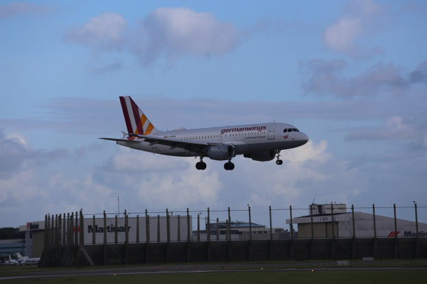 Germanwings Airbus A320-200 beim Abheben vom Frankfurt Airport, mit Gras darunter, einem Zaun im Vordergrund, Gebäuden und Bäumen im Hintergrund und einer bewölkten Himmel.
