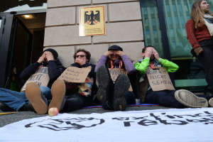 Eine Gruppe von Menschen sitzt vor einem Gebäude auf dem Boden und hält Schilder hoch, mit einer Fahne und einem Apfel auf der Straße und einer Frau auf der rechten Seite, die besorgt wirkt.