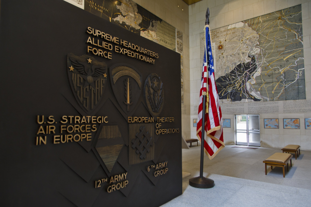 Eingang zum Hauptquartier der US-Strategischen Luftstreitkräfte in Europa mit einer amerikanischen Flagge, Text und Logos an der Wand, Bänken, einer Tafel, Fotorahmen, beleuchteter Decke und einer Tür im Hintergrund.