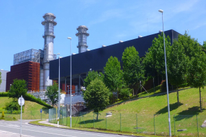 Ein großes Gebäude mit zahlreichen Fenstern, identifiziert als Kraftwerk durch die Beschriftung, umgeben von Straßeninfrastruktur, Fahrzeugen, Grünflächen und einem bewölkten Himmel.