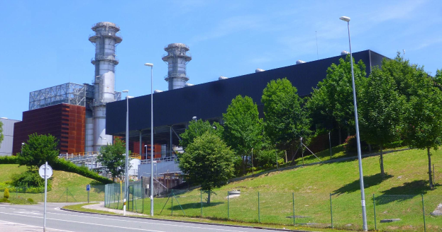 Ein großes Gebäude mit zahlreichen Fenstern, identifiziert als Kraftwerk durch die Beschriftung, umgeben von Straßeninfrastruktur, Fahrzeugen, Grünflächen und einem bewölkten Himmel.