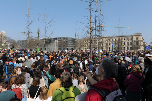 Eine große Menge Menschen mit Schildern und Taschen vor einem Gebäude mit Bäumen und einem klaren Himmel, was auf eine Klimawandel-Demonstration hinweist.