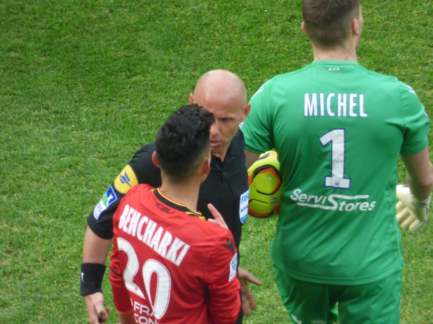 Zwei Fussballspieler und ein Schiedsrichter auf einem grasbedeckten Feld, wobei einer einen Ball hält.
