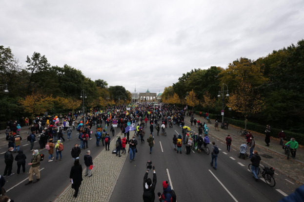 Eine große Gruppe von Menschen marschiert auf einer baumbestandenen Straße mit Laternenmasten, hält Kameras, mit einem Gebäude im Hintergrund und einem klaren Himmel darüber.