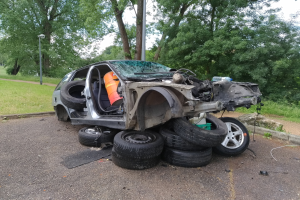 Ein umgestürzter Wagen am Straßenrand umgeben von Gras, Bäumen und Trümmern nach einem Unfall.