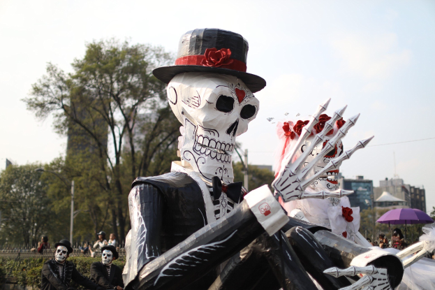 Ein Day of the Dead-Umzug in Mexiko-Stadt mit Menschen in Kostûmen mit Schirmen und skelettartigen Figuren vor einem Hintergrund aus Bâumen, Gebâuden und einem bewölkten Himmel.