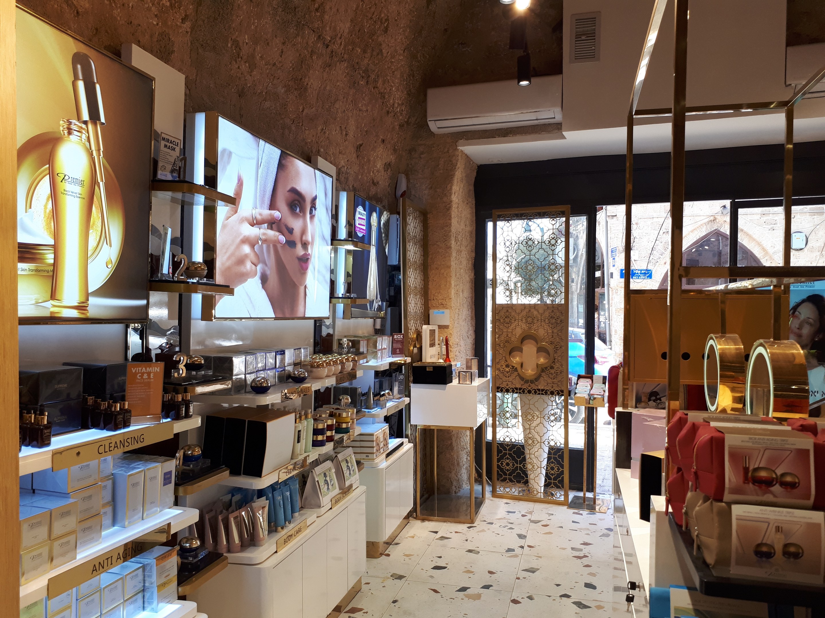 Ein gut beleuchteter Kosmetikgeschäftsinnenraum mit Regalen voller verschiedener Schönheitsprodukte, Wandbildschirmen, Deckenleuchten, einer Klimaanlage und Glastüren, die eine Person, ein Auto und ein Gebäude draußen zeigen.