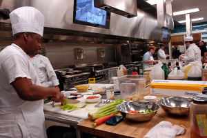 Eine Gruppe von Köchen in einer Küche, die Essen auf einem Schneidebrett zubereiten, mit einem Koch im Vordergrund in einer weißen Schürze und Mütze, der ein Messer hält, und umgeben von verschiedenen Utensilien, Schüsseln, Gemüse, einem Handy, Flaschen und anderen Gegenständen, mit einem Fernseher und Deckenlampen im Hintergrund.