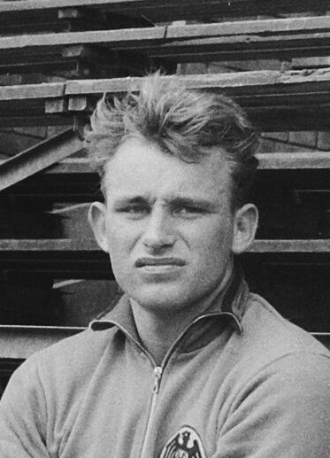 Schwarzes und weißes Foto eines Mannes in einer Jacke, der vor einem Stapel Holzplanken steht, der als legendärer deutscher Fußballspieler gilt.