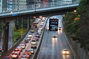 Eine belebte Stadtstraße mit starkem Verkehr, eine Brücke im Hintergrund und ein Banner mit Text in der Mitte der Straße; rechts sind Bäume, Pfosten, Laternen, Schilder und Gebäude zu sehen.