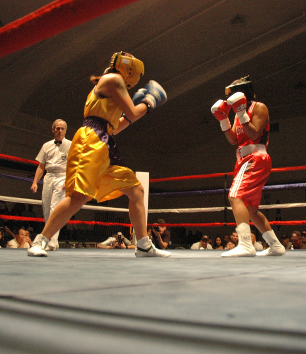 Zwei Boxer kämpfen in einem Ring mit einem Schiedsrichter im Hintergrund, umgeben von Fotografen und Zuschauern unter einer sichtbaren Decke.