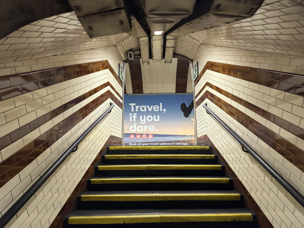 Eine Treppe in einer U-Bahn-Station mit einem Schild, auf dem "Reise, wenn du dich traust" steht, Geländer auf beiden Seiten und Stationwände im Hintergrund.