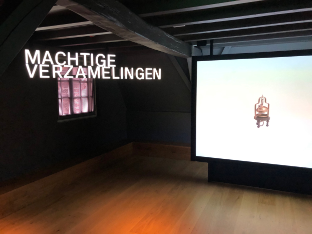 Installationsansicht der Ausstellung 'Machtige Verzamelingen' im Museum of Modern Art in Berlin, mit einem großen Bildschirm rechts, Text an der Wand dahinter und einem Fenster links.