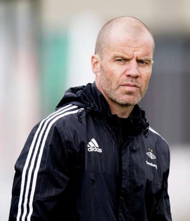 Ein kahlköpfiger Mann in einer schwarzen Jacke steht auf einem Fußballfeld, trägt eine marineblaue Jacke mit weißen Streifen und einem weißen Kragen und hat die Hände gefaltet, mit einem ernsten Gesichtsausdruck.