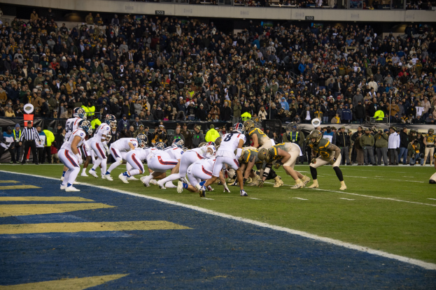 Ein Fußballspiel im Gange auf einem Stadionfeld mit Spielern in Sportbekleidung und Helmen, umgeben von Zuschauern und Stadionelementen im Hintergrund.
