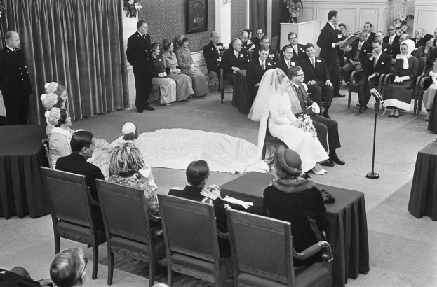 Schwarzes und weißes Foto einer Hochzeitszeremonie mit Menschen, die sitzen und stehen, einem Mann mit Mikrofon, einer Frau mit einem Blumenstrauß, einem Podium mit Mikrofon, Blumenschmuck, einem Vorhang, einem gerahmten Foto an der Wand und einer Tür im Hintergrund.