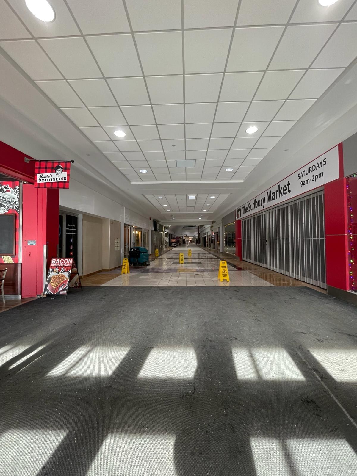 Innenansicht eines Einkaufszentrums mit Deckenbeleuchtung, einem Stuhl und einer Tafel, die "Sydbury Market - Vorher und Nachher" links zeigt, und verstreuten Gegenständen rechts und auf dem Boden.