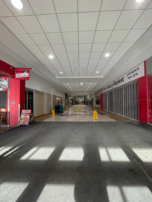 Innenansicht eines Einkaufszentrums mit Deckenbeleuchtung, einem Stuhl und einer Tafel, die "Sydbury Market - Vorher und Nachher" links zeigt, und verstreuten Gegenständen rechts und auf dem Boden.