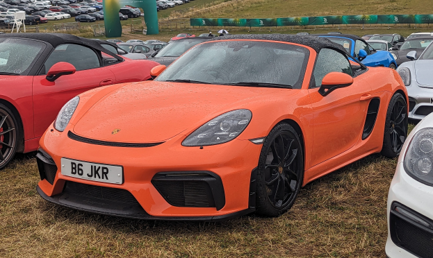 Roter Porsche 718 Boxster GTS auf dem Goodwood Festival of Speed mit Autos auf dem Rasen, einem Banner, Bäumen und einem klaren blauen Himmel.