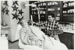 Ein Mann lächelt in einem Zahnarztstuhl, während ein anderer Mann ein Tablett neben ihm hält, mit einem Waschbecken, Sternen, einer Uhr und Regalen voller Flaschen im Hintergrund.