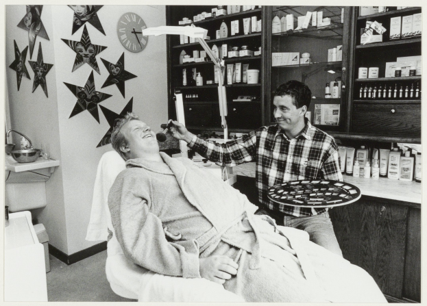 Ein Mann lächelt in einem Zahnarztstuhl, während ein anderer Mann ein Tablett neben ihm hält, mit einem Waschbecken, Sternen, einer Uhr und Regalen voller Flaschen im Hintergrund.