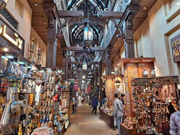 Ein belebter Dubai-Basar mit Menschen, die zwischen Regalen voller Gegenstände hindurchgehen, Wänden mit Text und einer Decke mit warmem Licht.