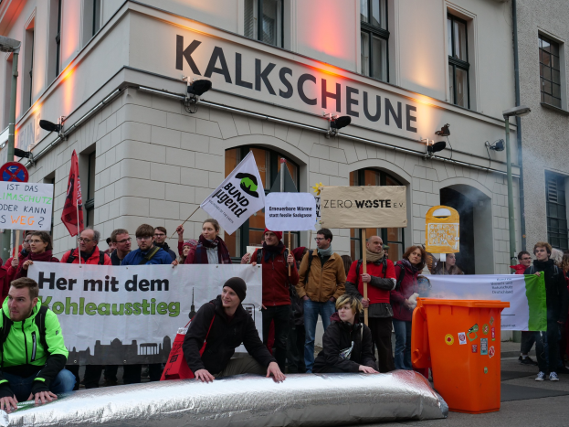Gruppe von Menschen mit Schildern und Plakaten vor einem Gebäude bei einer Demonstration in Deutschland, mit zwei Personen im Vordergrund und einem Müllcontainer auf der rechten Seite.