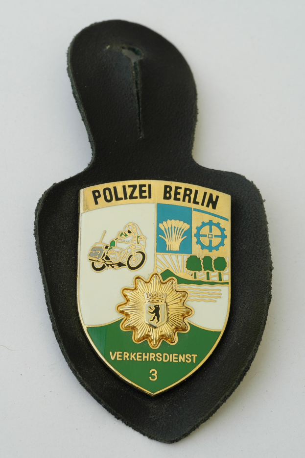 Ein schildförmiges Polizeibadgebnis mit "Polizei Berlin" in schwarzer fetter Schrift, das ein weißes Sternsymbol auf einem blauen Hintergrund mit einem weißen Rand zeigt, auf einer Fläche platziert.