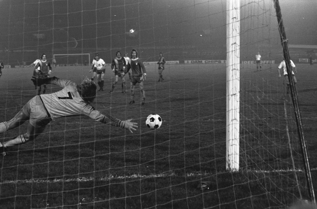 Ein Schwarz-Weiß-Foto eines Fußballspielers, der vor einem Tor einen Ball hält, umgeben von einer Gruppe von Menschen, die auf dem Boden stehen. Das Torpfosten ist auf der rechten Seite des Bildes zu sehen, sowie Werbetafeln und Lichter im Hintergrund.