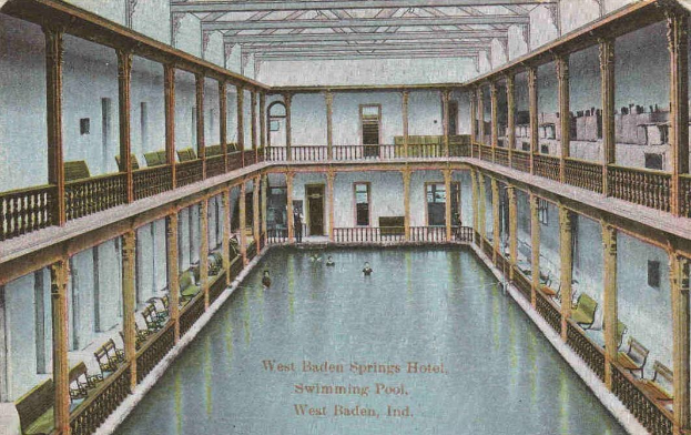 Schwarz-weiß-Postkarte eines Hallenbads im West Baden Springs Hotel mit schwimmenden Menschen, umgeben von architektonischen Details und Text am unteren Rand.