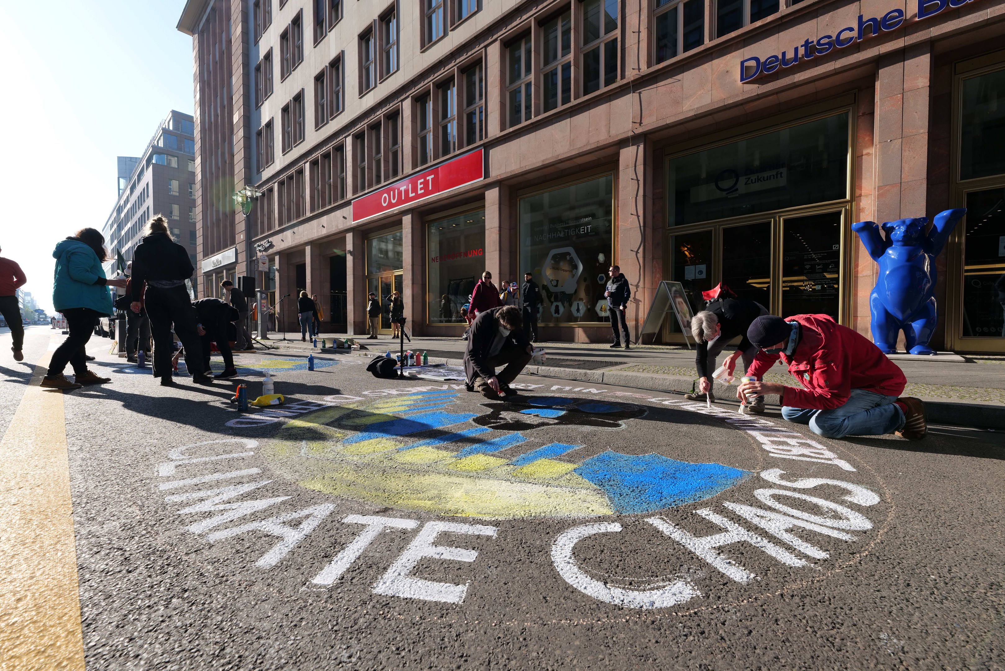 Menschen sitzen vor einem Gebäude mit Fenstern und Schildern auf dem Boden während einer Klimawandel-Demonstration in Berlin, umgeben von Flaschen und anderen Gegenständen, mit Bäumen und einem klaren blauen Himmel im Hintergrund.