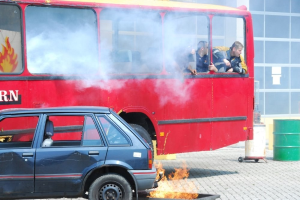 Ein roter Doppeldeckerbus mit Rauch, der aus ihm herauskommt, mit drei sichtbaren Passagieren, neben einem Auto geparkt, vor einem Gebäude mit Glasfenstern und einem Fass auf der rechten Seite.