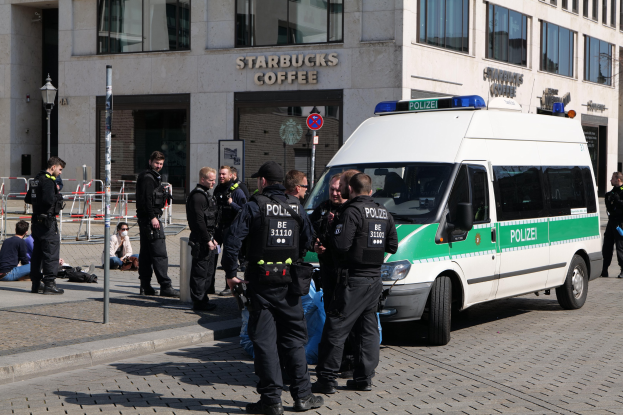 Polizeibeamte vor einem Starbucks-Café stehend mit einem Van auf der rechten Seite und Menschen auf der linken Seite sowie einem Gebäude, einem Schild, einem Laternenpfahl und einem Zaun im Hintergrund.