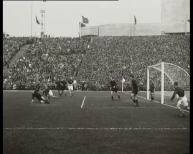 Schwarzes und weißes Foto des WM-Finales 1958 zwischen Manchester United und Liverpool, das Spieler auf dem Feld, ein Torpfosten auf der rechten Seite, Zuschauer in den Rängen und Fahnen im Hintergrund zeigt.