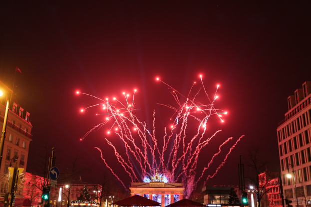 Eine belebte Stadtstraße an einem Silvesterabend in Berlin, mit Gebäuden, Bäumen, Laternen, Verkehrszeichen, Zeltplanen, Menschen und einem prächtigen Feuerwerk am Himmel.