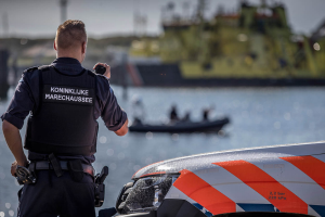 Ein Polizist in Uniform steht neben einem Polizeiauto in der Nähe eines Gewässers und hält ein Funkgerät in der Hand, im Hintergrund ist ein Boot mit ein paar Menschen auf dem Wasser zu sehen.