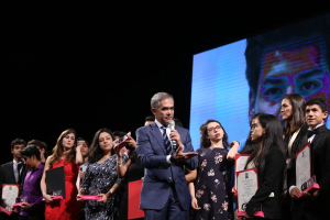Ein Mann im Anzug und mit Krawatte hält ein Mikrofon auf einer Bühne, umgeben von Menschen mit Auszeichnungen, mit einem sichtbaren Bildschirm im Hintergrund.