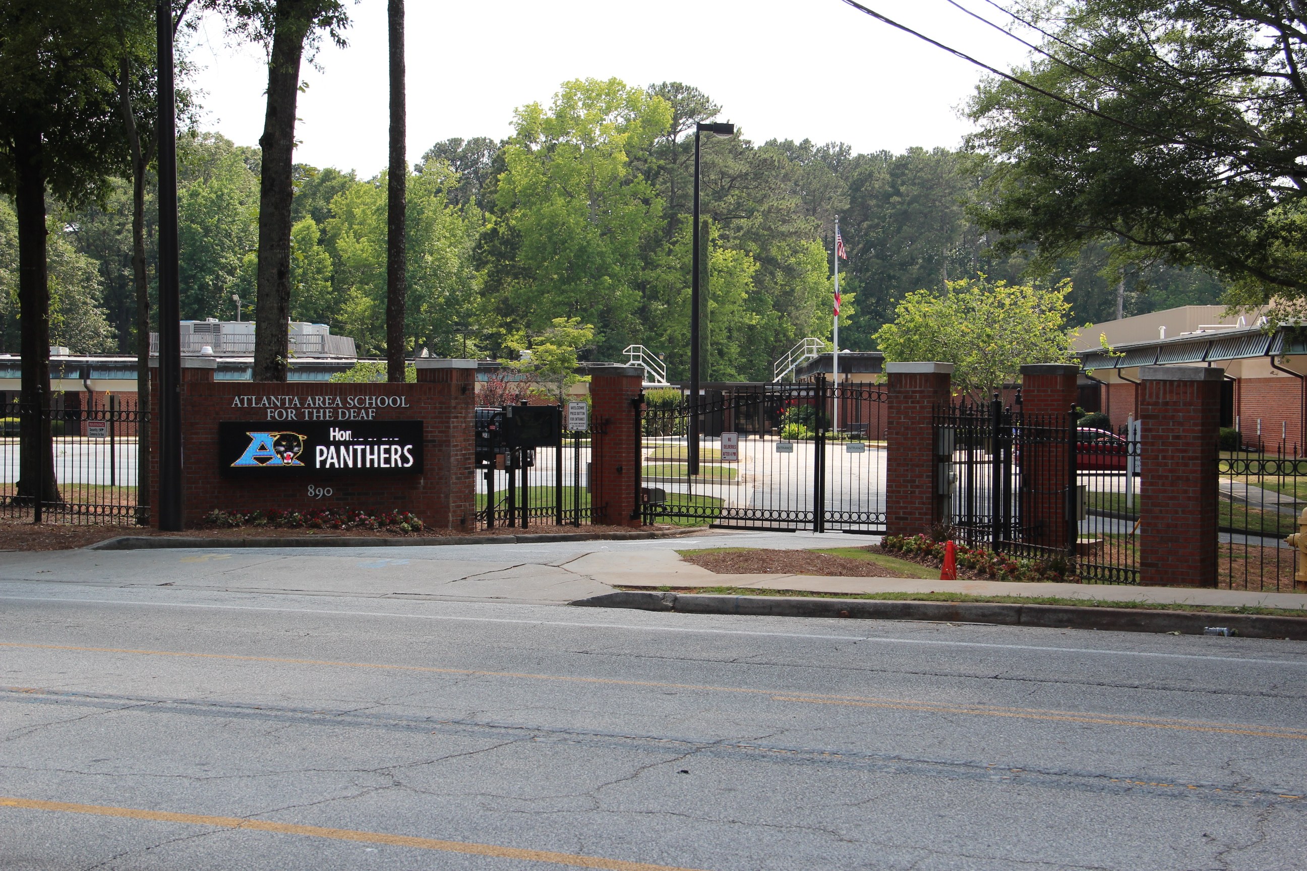 Schulbeginn mit einem Schild, umgeben von Bäumen, Straßenmöbeln, Fahrzeugen und Gebäuden unter einem klaren blauen Himmel.