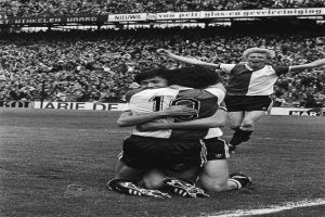 Drei Fussballspieler umarmen sich auf dem Boden beim Feiern eines Tores, mit jubelnder Menge und schwenkenden Fahnen im schwarz-weißen Hintergrund.