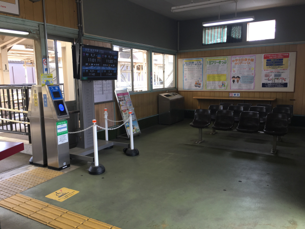 Ein belebter Bahnhofseingang mit Sitzgelegenheiten, einer Informations counter, Ticketautomaten, Kettenzaunpfählen, Informationsanzeigen, Wandplakaten, Deckenbeleuchtung und Geländern.