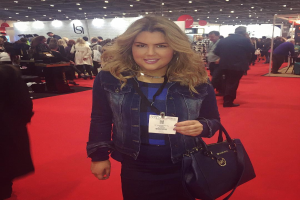 Eine Frau steht auf einem roten Teppich, lächelt und hält eine Handtasche in der Hand, mit Menschen, Lichtern und Schildern im Hintergrund bei einer Modenschau.