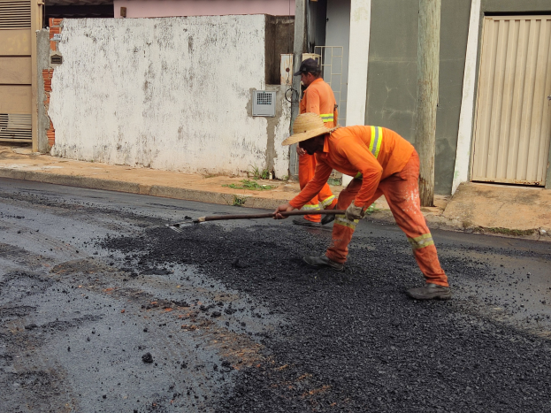 Zwei Männer in orangefarbenen Overalls und Hüten arbeiten an einer Straße, einer hält ein Objekt, mit Gebäuden und Läden im Hintergrund.