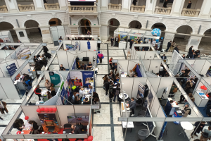Große Besuchermenge auf einer internationalen Fachmesse mit Ständen, Bannern, Tischen, Stühlen und Ballons in einem Raum mit Bögen, Säulen und einem Bildschirm im Hintergrund.