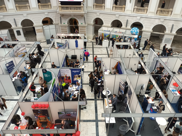 Große Besuchermenge auf einer internationalen Fachmesse mit Ständen, Bannern, Tischen, Stühlen und Ballons in einem Raum mit Bögen, Säulen und einem Bildschirm im Hintergrund.