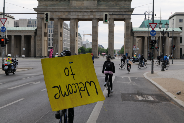 Eine Gruppe von Radfahrern mit Helmen fährt auf einer Straße am Brandenburger Tor in Berlin, Deutschland vorbei, wobei eine Person ein gelbes Schild hält, Lichtmasten, Verkehrszeichen, Gebäude, Bäume und einen klaren blauen Himmel im Hintergrund.