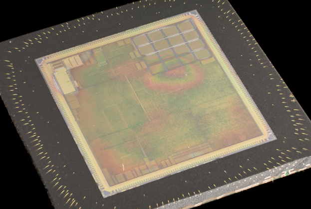 Nahaufnahme eines rechteckigen Halbleiterchips mit metallischem Glanz, umgeben von einem dünnen Metallrahmen und mit kleinen Löchern an seinen leicht erhöhten Rändern auf schwarzem Hintergrund.