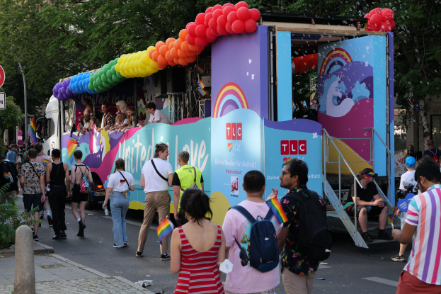 Eine Gruppe von Menschen geht neben einem Lastwagen mit bunten Luftballons die Straße entlang, mit Schildern an Pfosten an der Straße und Bäumen und Gebäuden im Hintergrund, was auf eine Pride-Parade in Paris hindeutet.