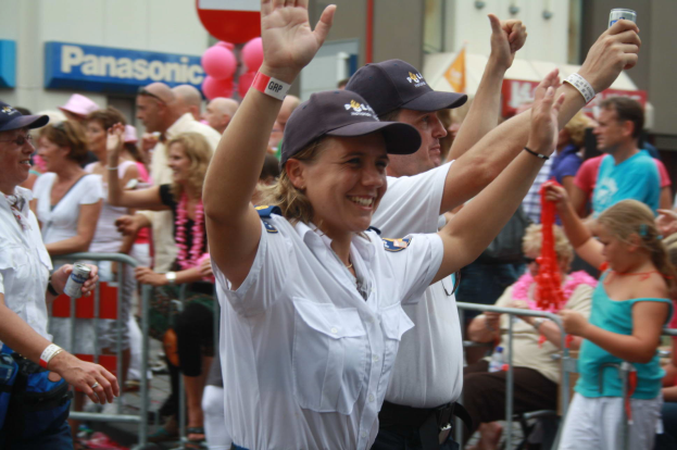 Eine Gruppe von Menschen, einige mit Mützen und Dosen tragend, steht und sitzt vor einer Menge, mit Geländern, Ballons und einem Gebäude mit Schildern im Hintergrund.
