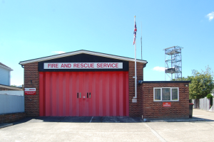 Feuerwehr- und Rettungsdienstgebäude mit roter Tür, Fenstern, einem Namensschild, einem Flaggenmast mit Flagge, einem Metallturm, einem Zaun, umliegenden Bäumen und einem bewölkten Himmel.