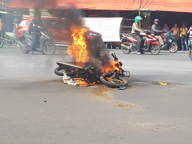 Ein in Flammen stehendes Motorrad am Straßenrand mit einem Auto und einem Van im Hintergrund, umgeben von Menschen, einige tragen Helme, mit einer Fahne und einem Zelt in der Szene.